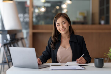 Asian businesswoman sitting happily with her laptop and takes notes intensely and smiles happily at her assignment.