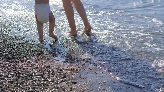 Close up view of mother helping one year old son in diaper to go onto the cold sea water on the Black sea beach with pebbles. Summer vacation, holiday, family trip. Sea without heavy waves