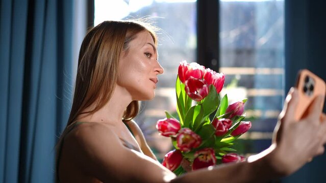 Side view charming young woman taking selfie with bouquet of flowers smiling. Positive happy Caucasian beautiful lady photographing with red tulips at home on holiday. Women's day and Valentines