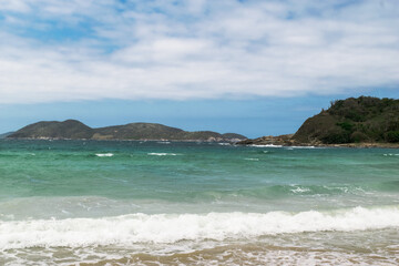 Linda praia das Conchas, próxima a cidade de Cabo Frio, com mar de águas verdes, céu com nuvens e montanhas ao fundo.