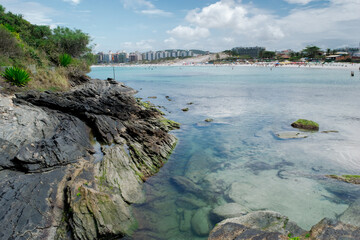 Lindas praias rochassas do forte em volta do Forte São Mateus em Cabo Frio, a água azul do mar correndo entre elas, algumas pessoas nas rochas e montanhas ao fundo.