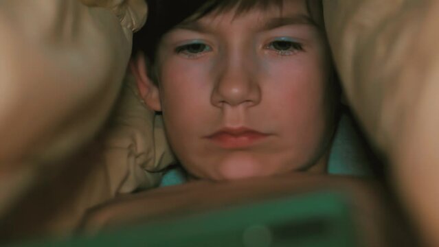 A child in bed under a blanket with a phone in his hands. Little boy watching video on smartphone in bed. The child looks at the phone emotionally. Selective soft focus.