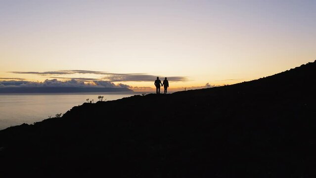 Aerial silhouette of a couple giving each other a high five celebrating successful climb on the mountain in beautiful sunset. Ocean background.