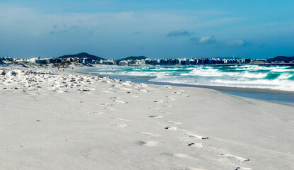 Orla da Praia das Dunas, com algumas dunas de areia, céu azul e a cidade de cabo frio ao fundo.
