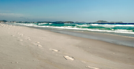 Orla da Praia das Dunas, com muitas pegadas na aréia, céu azul e muitas montanhas ao fundo.
