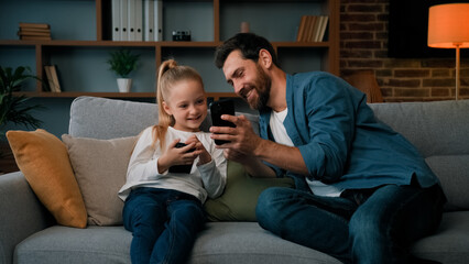 Caucasian man and little kid girl sit together at home hold modern phone browsing media content...