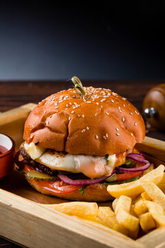 Beef burger with tomato, fried eggs, pickle, onion, lettuce, cheese sauce on a wooden board with french fries and ketchup.
