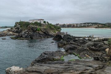 Praia do Forte, com o forte no topo de monte, mar, grandes rochas, céu com nuvens e a cidade de...