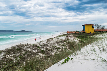 Fototapeta premium Vista da orla da Praia das Dunas, com lindo mar, dunas de areia branca, vegetação rasteira, construções litorâneas, céu azul e muitas montanhas ao fundo.