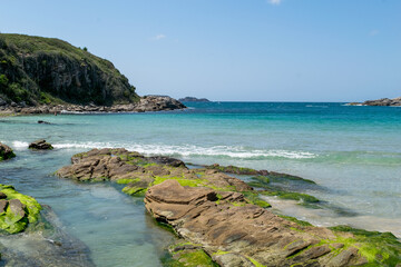 Vista da praia das Conchas, próxima a cidade de Cabo Frio, com mar azul em volta, vegetação rasteira, rochas com musgo verde e montanhas ao fundo.