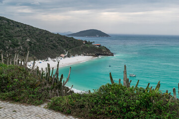 Obraz premium Vista de cima da linda Praia Arraial do Cabo Atalaia, com suas areias brancas, um lindo mar em volta, céu azul com nuvens, vegetação ao redor e cercada de grandes montanhas verdes.