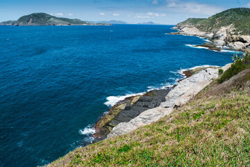Vista de cima da linda praia das Conchas, próxima a cidade de Cabo Frio, com muitas rochas a beira-mar, céu azul, mar com águas limpas e em tom de  azul, com montanhas ao fundo.