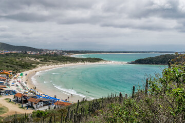 Vista de cima da linda praia das Conchas, próxima a cidade de Cabo Frio, com praias de areia branca, vegetação ao redor, céu azul, mar com águas limpas e em tom de verde e azul, com montanhas ao fundo