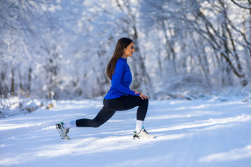 Fototapeta na wymiar Young sportswoman in perfect shape stretching leg in nature at snowy winter day.