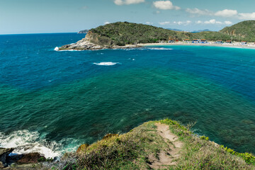 Vista de cima da linda praia das Conchas, próxima a cidade de Cabo Frio, com praias de areia branca, vegetação ao redor, céu azul, mar com águas limpas e em tom de verde e azul, com montanhas ao fundo