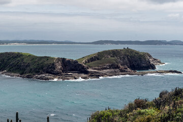 Vista de cima da praia das Conchas, pr&oacute;xima a cidade de Cabo Frio, mar azul, c&eacute;u com nuvens e muitas montanhas ao fundo.