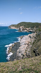 Vista de parte da linda praia das Conchas, com vegetação rasteira, um lindo mar azul em volta, grandes rochas, céu azul e montanhas ao fundo.
