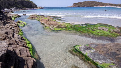 Vista de parte linda praia das Conchas, próxima a cidade de Cabo Frio, com mar azul em volta, vegetação rasteira, rochas com musgo verde e montanhas ao fundo.