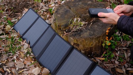 Hands plug in portable solar panel charging power bank by mutlipurpose cable, smartphone, watches, laptops nature background. Clean energy for using in camping or at home when NO electricity.
