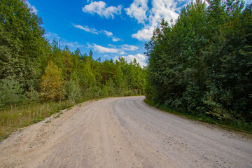Fototapeta na wymiar A gravel road curve in late summer or autumn with forest on the sides, blue cloudy sky and copy space on the trees