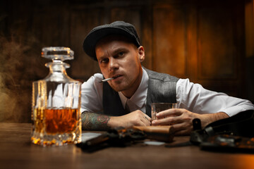Bold gangster sitting with cigarette at table with cards, pistol and decanter of whisky