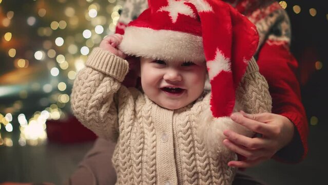 Mom puts a red Christmas hat on her baby. Family Spending Winter Evening at Home. Young Mother with Little Kid Son play. Slow Motion. Christmas, New Year, Winter Christmas Holiday Celebration.