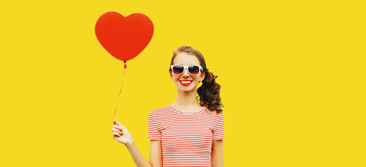Portrait of happy smiling girl with red heart shaped balloon wearing sunglasses isolated on yellow background