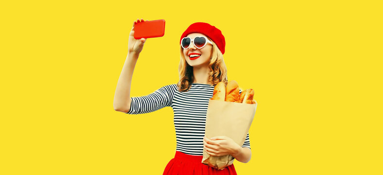 Portrait of happy smiling woman taking selfie with smartphone holding grocery shopping paper bag with long white loaf bread wearing red beret, heart shaped sunglasses isolated on yellow background