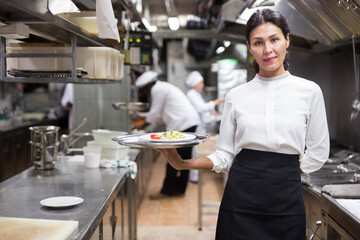 Fototapeta na wymiar portrait of happy female waitress standing in white kitchen in cafe