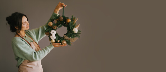 Millennial woman making Christmas wreath using natural pine branches and festive decorations in...