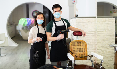 Portrait of male and female skilled hairstylist wearing protective face masks and gloves standing in modern hair studio, ready to work after coronavirus outbreak