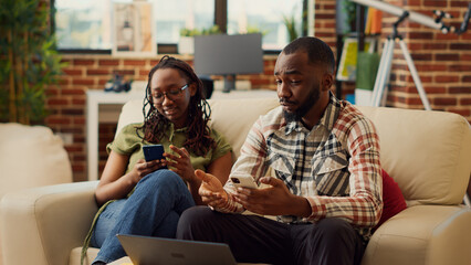 Obraz premium Young partners using mobile pohnes together on couch, browsing internet and scrolling through social media. People in love laughing at digital gadget, enjoying leisure activity relaxing.