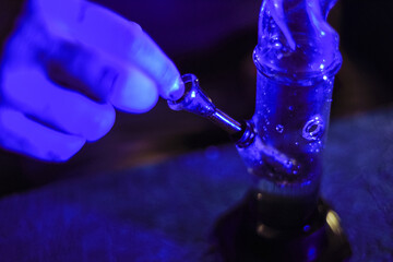 Male hands clog a bong with marijuana. Drugs and drug addict. The man smokes weed. Neon blue light. CBD or THC.