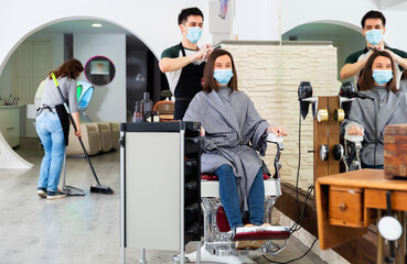 Young cheerful positive glad male professional hairdresser in mask cutting woman's hair in hairdressing salon