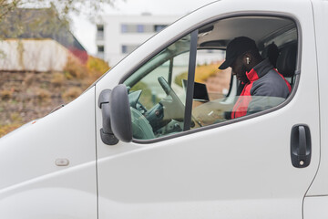 Fototapeta premium Hard-working, focused ethnic male courier during his shift. African-American man in black baseball hat sitting behind the wheel of white delivery van and checking things on his clipboard. High quality