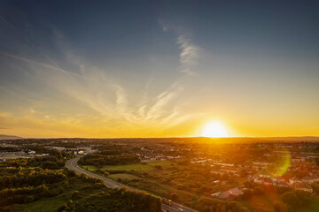 Fototapeta na wymiar Road in town by a green park and residential area with homes at warm sunset. Sunset time. Stunning nature scenery in the background. Galway city, Ireland. Aerial view. City transportation system.