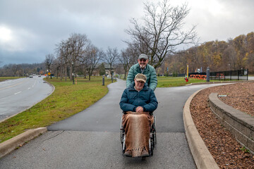 An older gay senior man, pushes his husband in a wheelchair down an incline path on a cool autumn day.  The man in the wheelchair has a blanket on his lap and both men are wearing winter jackets.
