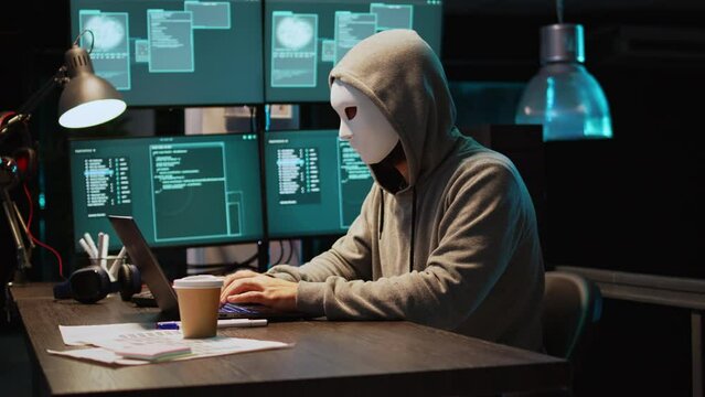 Cyber scammer wearing mask and hood to hack computer system, breaking into servers to steal big data. Masked man looking dangerous and scary, impostor creating security malware. Handheld shot.