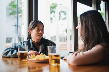 Two girl friends speaking with each other while having a snack and a beer in a bar