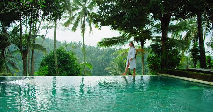 Woman enjoying summer vacation in luxury tourist resort amazing rice terraces near to swimming pool in sunny morning Bali. Girl walking in bathrobe by poolside, palm forest jungle on tropical island.