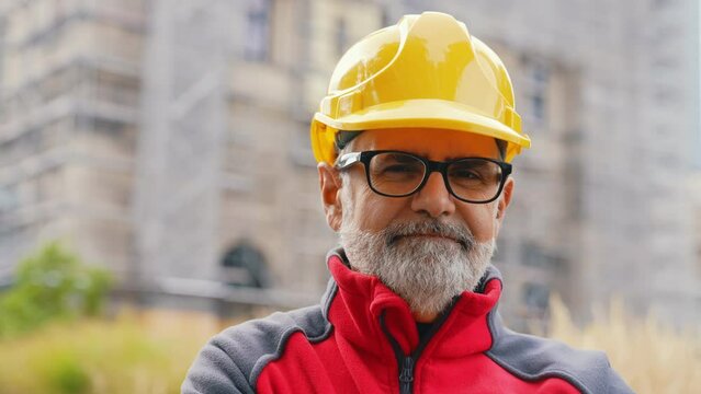 Portrait of a smiling, confident, grey-haired, middle-aged professional worker in uniform wearing a helmet and eyeglasses, standing in front of the construction site, looking at the camera. High
