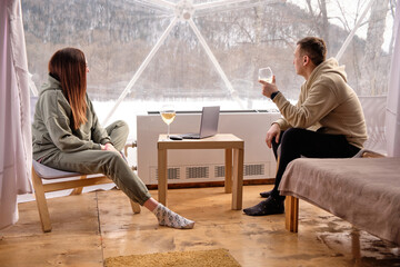 A guy and a girl with glasses of white wine look out the window in glamping. In winter, outdoors in a tent. The computer is on the table.