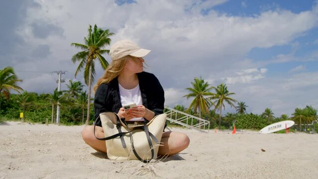 Young girl sitting on sand at the beach playing with her hair. Woman waiting at windy beach, with phone. Teen relaxing at the shore, between tall palm trees. Tall green palm trees background. 4k