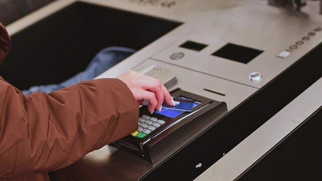 The hands of a young caucasian girl with a beautiful light manicure in a brown raincoat make a payment with a bank card in the self-service section of the store, side view close-up with slow motion, s