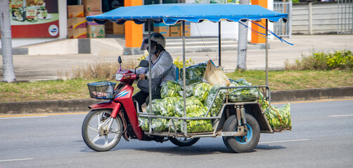 A woman rides a motorcycle with a sidecar loaded with plastic bags full of cucumbers, Thailand