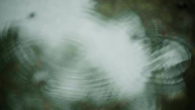 梅雨、砂利の地面の水たまりに雨の水滴が落ちて波紋を残すスローモーション