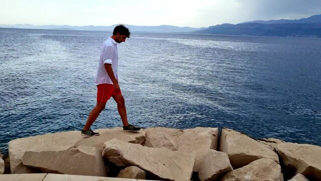 Young Man Walking Carefully On Rocks In Postira, In Front Of Blue Seascape, Croatia
