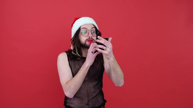 Young sexy transsexual our androgynous man in a Santa hat paints lips isolated on red background. People lifestyle fashion lgbtq concept