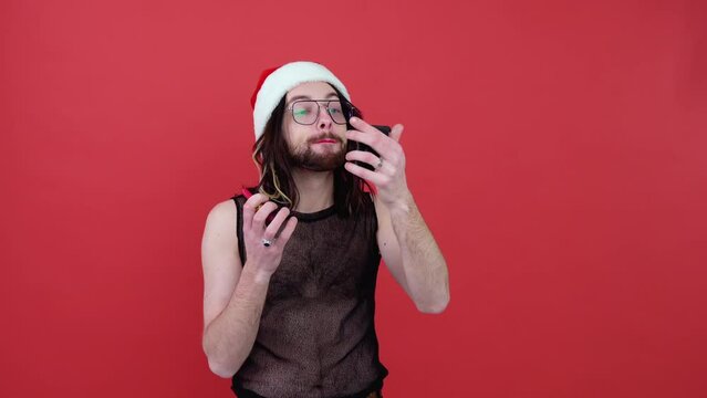 Young sexy gay man in a Santa hat paints lips isolated on red background. People lifestyle fashion lgbtq concept