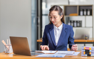 Brown haired wearing light blue jacket smiling Asian woman work with document laptop in office, doing planning analyzing the financial report, business plan investment, finance analysis concept.